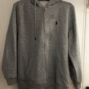 Säljer en snygg och bekväm grå hoodie från Ralph Lauren. Den har en dragkedja framtill och en klassisk logga på bröstet. Perfekt för kyliga dagar eller som ett stilrent lager. Passar både till vardags och avslappnade tillfällen.