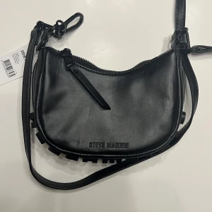 Svart axelväska från Steve Madden - Snygg svart axelväska från Steve Madden i syntetmaterial. Väskan har en dragkedja upptill och ett coolt logotypmönster på sidan. Perfekt för både vardag och fest! Helt oanvänd.