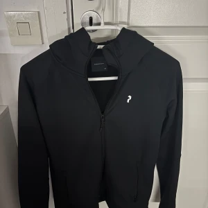 Svart hoodie från Peak Performance - Säljer en svart hoodie från Peak Performance i mycket bra skick. Den har en dragkedja framtill och en snygg logga på bröstet. På baksidan står det 'PEAK PERFORMANCE' i vitt. Perfekt för höst och vår, och den är superbekväm att ha på sig. Passar både till vardags och träning!