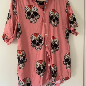 Rosa skjorta med dödskallemönster från Dickies - Säljer en cool rosa skjorta från Dickies med ett unikt dödskallemönster. Skjortan har korta ärmar och knappar framtill. Perfekt för en avslappnad stil med en edgy touch. Passar bra till både vardag och fest!
