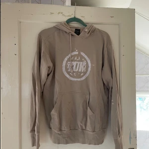 Beige hoodie från TDK - Säljer en beige hoodie från TDK i storlek M. Den har en stor ficka fram och en cool logga i vitt på bröstet. Perfekt för höst och vinter när du vill hålla dig varm och bekväm. Den är i bra skick och redo för nya äventyr!