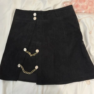 Svart kjol med kedjedetaljer - Säljer en supercool svart kjol med snygga guldkedjor och knappar framtill. Kjolen har en stilren design med en liten slits som ger den en extra edge. Perfekt för en utekväll eller fest! 🖤