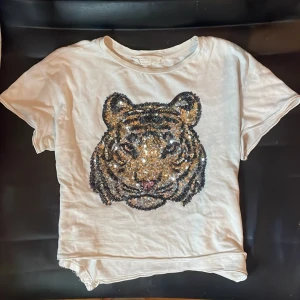Vit t-shirt med paljett-tiger från Zara Girls - Säljer en supercool vit t-shirt från Zara Girls med en stor paljett-tiger på framsidan. Den har korta ärmar och ett tryck på baksidan med texten 'FRIENDS MAKE THE ADVENTURE SO MUCH BETTER'. Perfekt för en avslappnad och stilren look! 🐯✨