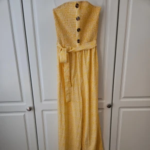 Gul axelbandslös klänning med knappar -  Nytt Säljer en superfin gul jumpsuit  med ett sött vitt mönster. Jumpsuiten har en smockad överdel med stora dekorativa knappar och ett knytband i midjan för en perfekt passform. 🌼