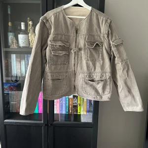 Utility Jacket Brown - Beige/brun jeansjacka från Djerf Avenue i absolut superskick! Har inga tecken på användning mer än att tvättlappen är en aning blek i färgen. Har tvättats i tvättmaskin men enligt tvättlappen. 100% bomull   Ärmarna kan tas av så det blir en väst. Stä