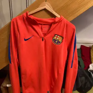 Säljer en snygg röd Nike-tröja med Barcelona-logga på bröstet. Tröjan har en dragkedja vid halsen och är långärmad. Perfekt för fotbollsträning eller som en casual sportig look. Den är i bra skick och har blå detaljer på ärmarna.