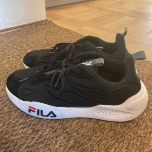 Svarta sneakers Fila - Säljer ett par snygga svarta Fila sneakers i nyskick. De har en vit sula med Fila-loggan i rött och blått på sidan. Skorna har snörning och en bekväm passform, perfekta för både vardag och träning. De är stilrena och lätta att matcha med olika outfits.