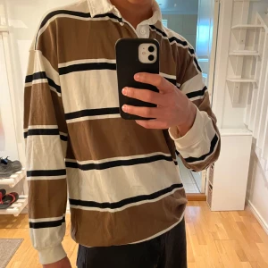 Randig pikétröja - Säljer en snygg och bekväm randig pikétröja från H&M i oversized fit. Tröjan har breda ränder i brunt, vitt och svart och är perfekt för en avslappnad stil. Den har långa ärmar och en klassisk krage. Perfekt för både vardag och chillhäng!