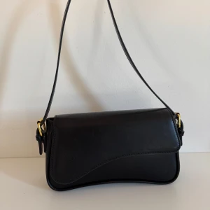 Svart axelväska i skinn - Säljer en stilren svart axelväska. Den har en enkel och elegant design med en justerbar axelrem och guldfärgade detaljer. Perfekt för både vardag och fest!