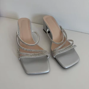 Silvriga sandaler med klack och glittriga remmar - Säljer ett par eleganta silvriga sandaler med fyrkantig tå och klack. De har flera tunna, glittriga remmar som ger en lyxig känsla. Perfekta för festliga tillfällen eller en utekväll. Skorna är i nyskick och redo att bäras av någon som vill glänsa!