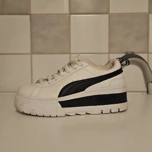 Vita sneakers från Puma - Säljer ett par snygga vita sneakers från Puma i storlek 38. Skorna har en cool svart detalj på sidan och en rejäl sula som ger extra komfort. De är i nyskick och perfekta för både vardag och fest. Skorna har snörning och är tillverkade i syntetmaterial.