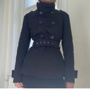 Svart kappa med bälte - Säljer en stilren svart kappa med dubbelknäppning och bälte i midjan. Jackan har långa ärmar och en hög krage som ger en elegant look. Perfekt för höst och vår!