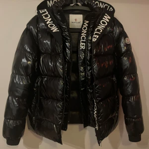 Svart dunjacka från Moncler - Säljer en snygg svart dunjacka från Moncler i bra skick. Jackan har en glansig finish och Moncler-logga på vänster ärm. Den är långärmad med dragkedja och har coola Moncler-band längs med dragkedjan. Perfekt för vintern! Den har två dragkedjor varav ena låter märket MONCLER synas längst med. Den yttre dragkedjan har en liten skada längst ner, påverkar inte funktionen märkbart. Serien på insidan har lossnat något. 