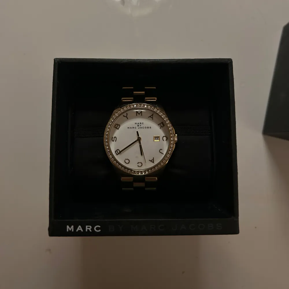 Säljer en elegant guldfärgad klocka från Marc by Marc Jacobs. Den har en vit urtavla med glittriga detaljer runt boetten och tydliga bokstäver som markerar timmarna. Armbandet är i metall och matchar den lyxiga stilen. Perfekt för både vardag och fest! ✨finns även extra länk . Accessoarer.