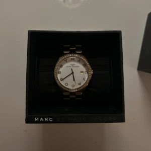 Guldfärgad klocka från Marc by Marc Jacobs - Säljer en elegant guldfärgad klocka från Marc by Marc Jacobs. Den har en vit urtavla med glittriga detaljer runt boetten och tydliga bokstäver som markerar timmarna. Armbandet är i metall och matchar den lyxiga stilen. Perfekt för både vardag och fest! ✨finns även extra länk 