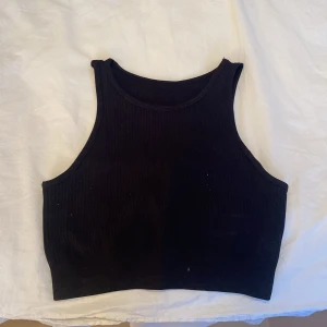 Svart ribbad linnetopp - Snygg svart ribbad linnetopp i ett stretchigt material. Perfekt för sommardagar eller som ett lager under en jacka. Toppen är ärmlös och har en croppad passform som ger en trendig look. Är en sport topp