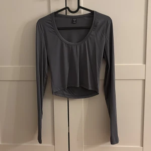 Grå långärmad topp - Snygg grå långärmad topp i syntetmaterial. Den har en rundad halsringning och är croppad, vilket ger en modern look. Perfekt för både vardag och festliga tillfällen. Toppen är i mycket bra skick och passar perfekt till jeans eller kjol.
