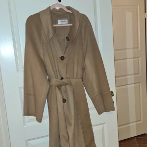 Beige kappa från Silk Maison - Säljer en elegant beige och lyxig kappa från Silk Maison i nyskick oanvänd. Den har långa ärmar och knäpps med stora knappar framtill. Kappan har ett bälte i midjan för en snygg passform. Perfekt för höst och vår! 100% wool. Slutsåld men nypris är ca 499 dollar