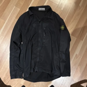 Svart jacka från Stone Island - Säljer en svart jacka från Stone Island i mycket bra skick. Den har en dragkedja framtill och en ikonisk logga på ärmen. Perfekt för höst och vår, med en stilren och modern look. Passar både till vardags och fest. Storleken är inte specificerad, men den ser ut att ha en normal passform.(köpt på johnells) nypris 5,2