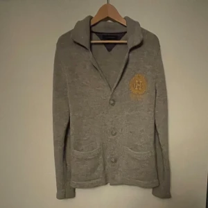 Grå kofta från Tommy Hilfiger - Säljer en snygg grå kofta från Tommy Hilfiger i storlek S. Den har en broderad logga på bröstet och knappar framtill. Perfekt för höst och vinter, med långa ärmar och praktiska fickor. Supermysig och stilren för alla tillfällen! 🍂 Är 180 och passar perfekt så lite stor i storleken! Pris går att diskuteras, skriv vid funderingar 🧐 