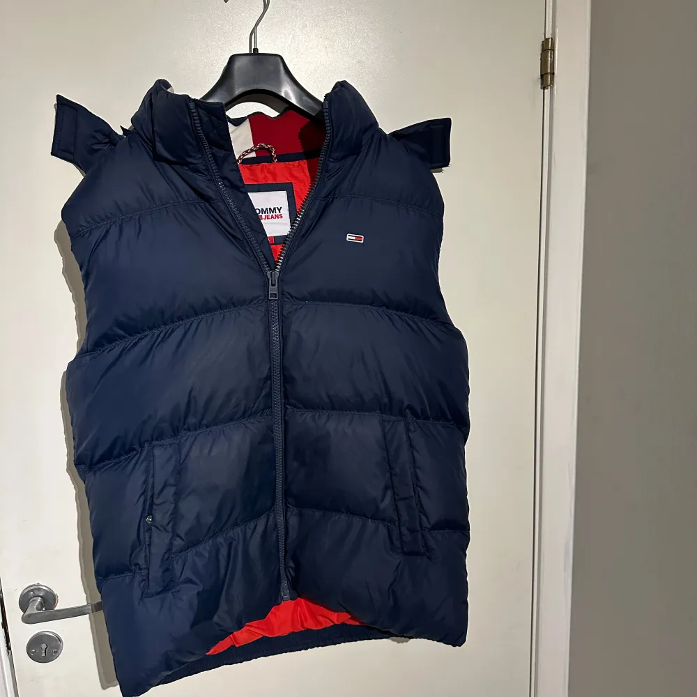 Säljer en snygg mörkblå dunväst från Tommy Jeans i storlek L. Västen har en dragkedja och en cool röd insida som ger en fin kontrast. Perfekt för höst och vinter, håller dig varm och stilren. Passar både till vardags och lite mer avslappnade tillfällen.. Jackor.