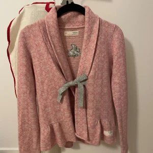 Rosa kofta från Odd Molly - Superfin rosa kofta från Odd Molly i ett mjukt och mysigt material. Den har en söt grå rosett framtill och ett broderat mönster på ryggen. Perfekt för höst och vår när du vill ha något bekvämt men ändå stiligt. Koftan har långa ärmar och en volangdetalj vid nederkanten.