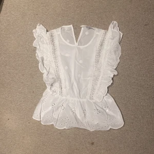 Vit blus med spetsdetaljer - Säljer en superfin vit blus med volanger och spetsdetaljer. Den har en söt broderad design längst ner och är ärmlös, perfekt för sommardagar. Blusen är i mycket bra skick och ger en romantisk och fräsch look.