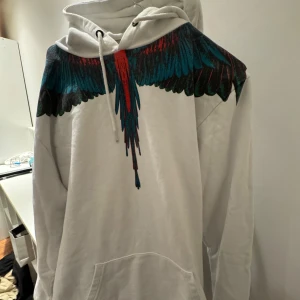 Vit hoodie med färgglatt fågel marcelo burlon - Säljer en snygg vit hoodie med ett coolt fågeltryck i blått, rött och svart över axlarna. Perfekt för att sticka ut i mängden! Den har en klassisk passform med känguruficka och justerbar huva. Perfekt för höst och vår.