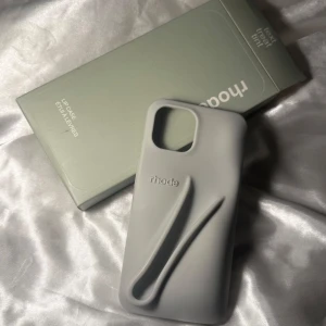 Grått mobilskal från Rhode - Säljer ett stilrent grått mobilskal från Rhode. Skalet har en unik design med en inbyggd hållare för läppbalsam, perfekt för att ha allt på ett ställe. Passar iPhone 14 och kanske 15 och är gjort av slitstarkt material. Perfekt för dig som vill ha både stil och funktion i ett.
