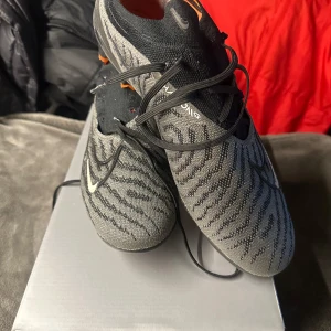 Nike Phantom fotbollsskor - Säljer ett par Nike Phantom elite fotbollsskor i grått och svart med orange detaljer. De är i storlek 44,5 och i väldigt bra skick. Förutom att ena skon saknar skosnören, men de är väldigt lätt att lösa👍