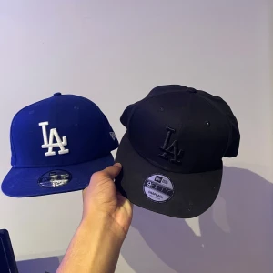 Två kepsar från New Era - Säljer två snygga kepsar från New Era. Den ena är blå med vit broderad logga och den andra är helsvart med svart broderad logga. Perfekta för både vardag och sportiga aktiviteter. Justerbar passform för bästa komfort. Har nästan aldrig använt de. För att jag har 5 andra i andra färger som jag gillar mer. Vanligt priss för dessa nya är 1000kr