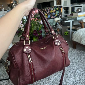 Vinröd skinnväska från Zadig & Voltaire - Säljer en snygg vinröd skinnväska från Zadig & Voltaire. Väskan har två praktiska dragkedjor framtill och en elegant logga i guld. Den saknar en guld platta på höger sida. Köpt begagnad från Vestaire för 2900kr för 1 år sen🥳