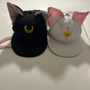 Sailor moon keps med kattöron - Två kepsar med söta kattöron och en gul måne framtill. En är svart och den andra är vit, båda med rosa insida på öronen.
