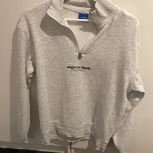 Säljer en stilren grå sweatshirt från Jack & Jones med halv dragkedja och texten 'Originals Studio' på bröstet. Perfekt för en avslappnad look. Tröjan har långa ärmar och är i mycket bra skick. Passar både till vardags och chillkvällar.