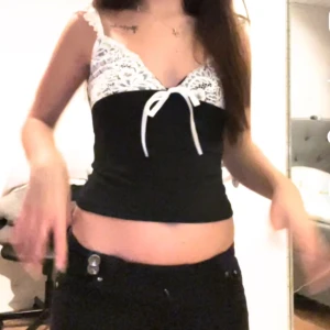 Svart och vit linne med spetsdetaljer - Snygg svart linne med vita spetsdetaljer och en söt knytning framtill. Perfekt för sommardagar eller en utekväll. Den ärmlösa designen och den croppade längden ger en trendig look.