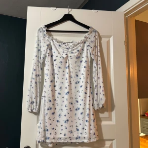 Vit blommig klänning - Superfin vit klänning med blå blommor, perfekt för vår och sommar! Den har långa ärmar och en söt rynkad detalj vid halsen. Materialet känns mjukt och bekvämt, perfekt för en dag ute i solen eller en mysig fika. Passar både till vardags och festligare tillfällen.