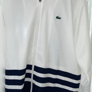 Vit och marinblå jacka från Lacoste - Säljer en snygg vit och marinblå jacka från Lacoste. Jackan har dragkedja och ett coolt randigt mönster längst ner. Den har långa ärmar och det klassiska Lacoste-loggan på bröstet. Perfekt för vår och höst!