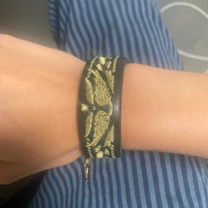 Svart och guld armband med broderi - Snyggt armband i svart med guldbroderi. Det har ett justerbart band och en unik design som passar perfekt för både vardag och fest. Broderiet ger en elegant touch och gör det till ett riktigt statement-smycke.