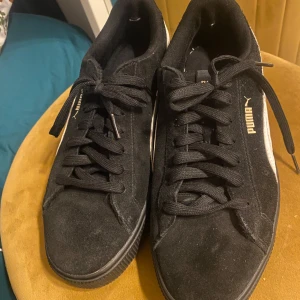 Svarta sneakers från Puma - Säljer ett par snygga svarta sneakers från Puma i bra skick. De är gjorda av mocka och har klassisk snörning. Perfekta för vardagsbruk och passar till nästan allt. Sulan är i bra skick och de har en stilren design med Pumas logga på sidan.