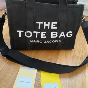 Svart tote bag från Marc Jacobs - Säljer en snygg svart tote bag från Marc Jacobs. Väskan har ett stilrent tryck med texten 'THE TOTE BAG' och är perfekt för vardagsbruk. Den har en praktisk dragkedja upptill och en justerbar axelrem för bekväm användning. Insidan är rymlig med flera fack för organisering. Perfekt för både skola och jobb!