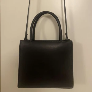 Svart axelväska från H&M - Snygg och stilren svart axelväska från H&M i syntetmaterial. Perfekt för både vardag och fest. Väskan har en enkel design med en lång axelrem och två handtag, vilket gör den lätt att bära på olika sätt. Passar till alla outfits och är ett måste i garderoben!