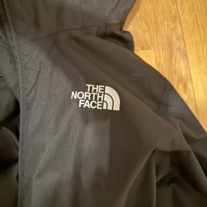 Svart vindjacka från The North Face - Säljer en svart vindjacka från The North Face i mycket bra skick. Jackan har en dragkedja framtill och är perfekt för höst och vår. Den är tillverkad i ett slitstarkt syntetmaterial och har långa ärmar. Perfekt för utomhusaktiviteter och vardagsbruk.