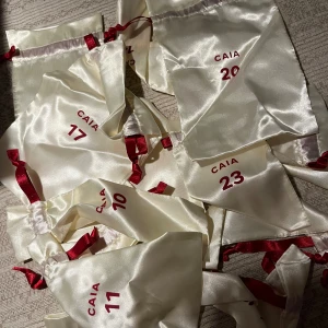 Vita satinpåsar från CAIA - Säljer ett gäng vita satinpåsar från CAIA med röda siffror och band. Perfekta för att förvara smink eller småsaker. De är numrerade, vilket gör dem idealiska för en adventskalender eller liknande. Superfina och praktiska!
