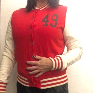 Röd och vit jacka från GANT - Säljer en snygg röd och vit jacka från GANT i storlek S. Den har ett coolt tryck med siffran 49 på framsidan och texten 'RUGGER' på baksidan. Jackan har långa ärmar och stängs med knappar. Perfekt för höst och vår!