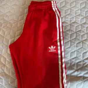 Säljer ett par snygga röda träningsbyxor från Adidas med klassiska vita ränder längs sidorna. Byxorna har en elastisk midja för extra komfort och är perfekta för både träning och chill. Adidas-loggan är broderad på sidan. Passar perfekt för dig som vill ha en sportig look!