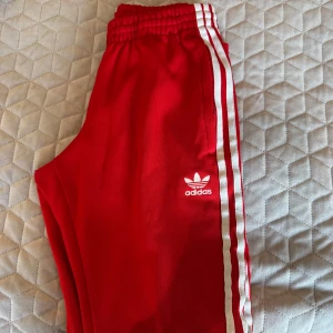 Röda träningsbyxor från Adidas - Säljer ett par snygga röda träningsbyxor från Adidas med klassiska vita ränder längs sidorna. Byxorna har en elastisk midja för extra komfort och är perfekta för både träning och chill. Adidas-loggan är broderad på sidan. Passar perfekt för dig som vill ha en sportig look!