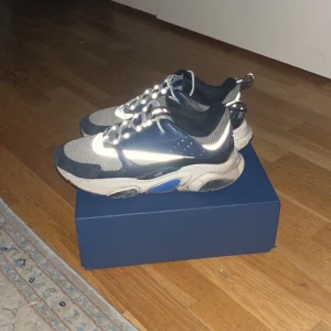 Dior B22 Navy - Säljer ett par stilrena sneakers från Dior i mycket bra skick. Skorna är i en kombination av grått och blått med vita detaljer och har en modern design. skorna är använda men har ändå mycket att leva för, fick i födelsedagspresent i sommar. Möts upp eller fraktar Östermalm 📍 passar 44 några sprickor i Sulan men de inget som inte kan fixas, allt kan tvättas och fixas på en skomakare 