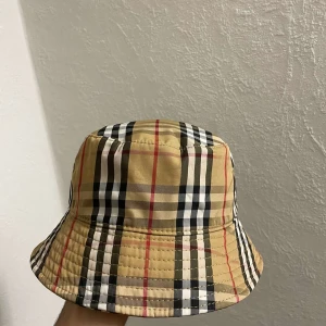 Burberry bucket hat - Snygg bucket hat med klassiskt burberry mönster i beige, svart, vit och röd. Perfekt för att ge din outfit en stilren touch. Hatten är aldrig använd och passar perfekt för sommarens alla äventyr.