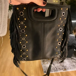 Svart axelväska med fransar och nitar - Maje väska,Säljer en supercool svart axelväska i läder. Den har fransar på sidorna och är dekorerad med nitar och ringar i guld och silver. Använd och tappat lite fransar på baksidan men inget man tänker på.