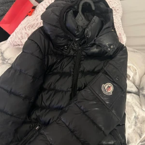 Svart dunjacka från Moncler - Säljer en svart dunjacka från Moncler. Den är varm och perfekt för vintern med en snygg glansig finish. Jackan har en dragkedja och en logga på ärmen. Det finns en liten skada vid ärmslutet som behöver fixas. Perfekt för dig som vill ha stil och komfort i kylan!